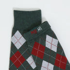 A close-up of a folded knit garment, likely a sweater or scarf, in dark green with an argyle pattern in red and white, featuring a small red and white embroidered logo on the cuff.