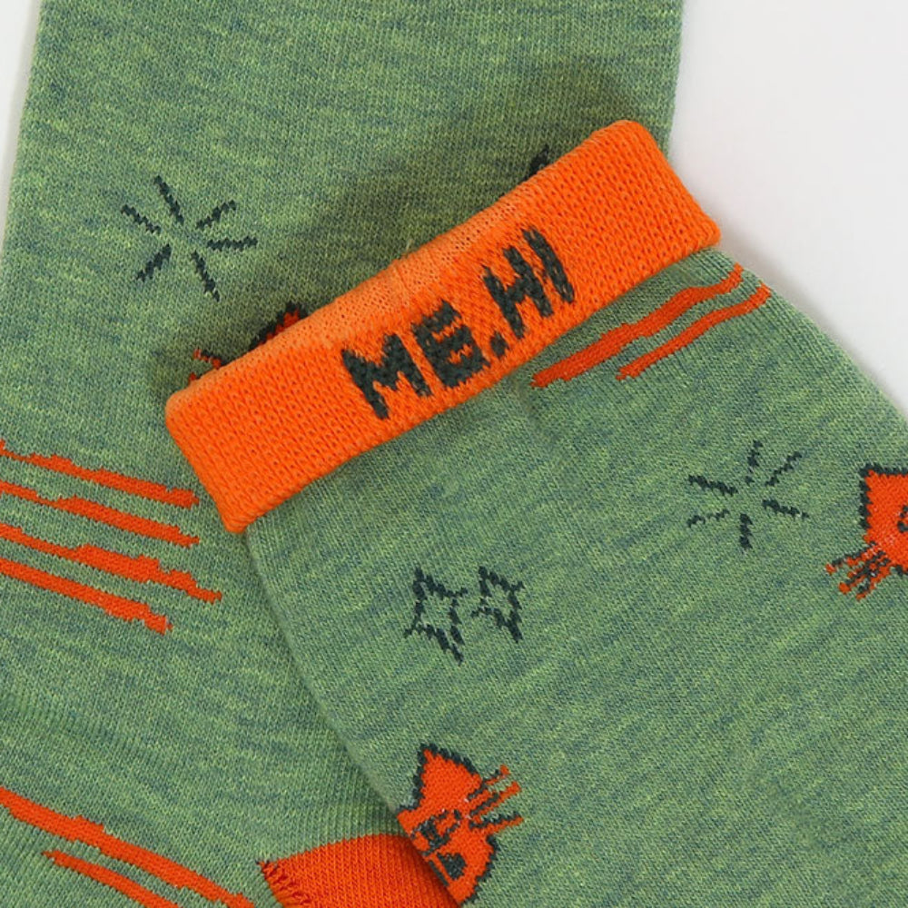 A close-up of a green knit sock with an orange cuff featuring the text 'ME, HI' in black. The sock has orange and black cat-themed graphics, including a cat's head, orange stripes, and black starburst shapes, shown against a plain white background.