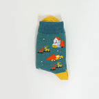 A single teal sock with a whimsical winter scene print, featuring a house, reindeer, and a red car, with yellow accents at the top and bottom, displayed on a clean white background.