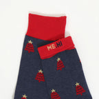 A pair of navy blue socks with a red ribbed cuff and a repeating pattern of red Christmas trees with gold stars. One sock is folded over the other, showing the red cuff with the text 'ME.