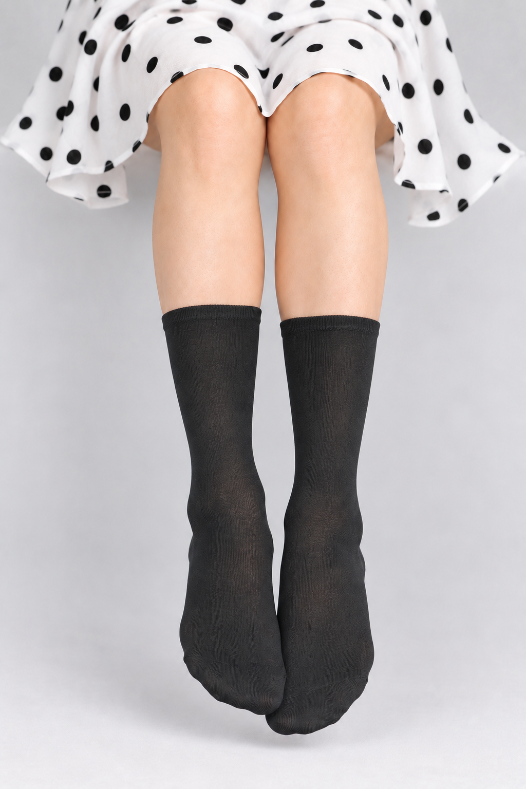 A person wearing black crew socks, shown from the waist down, with a white and black polka dot skirt visible at the top against a light gray studio background.