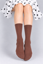 A person seated wearing a white dress with black polka dots and brown knee-high socks against a light gray studio background.