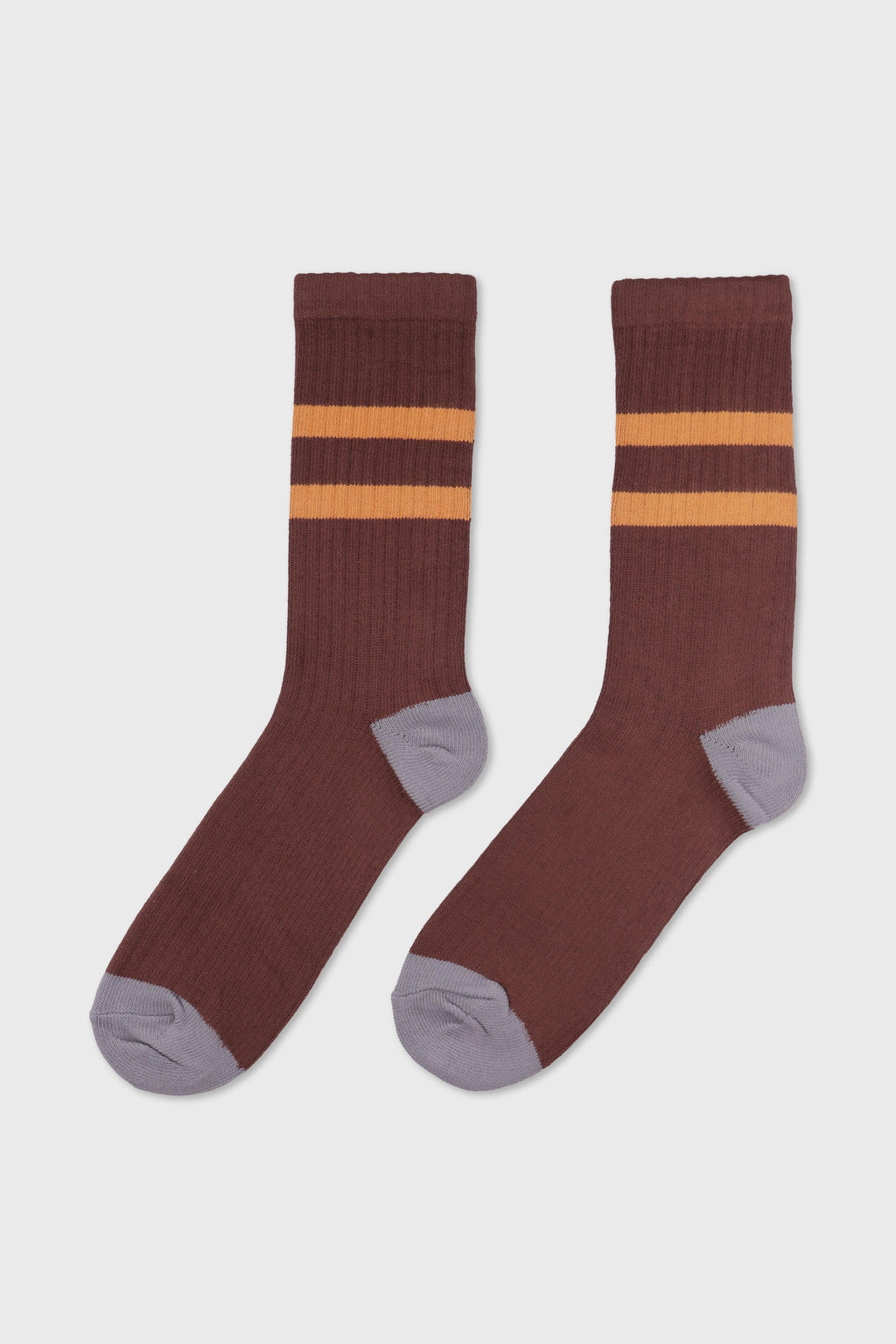 A pair of ribbed crew socks in a dark brown color, featuring two horizontal mustard yellow stripes near the cuff and contrasting light gray heels, toes, and soles, displayed against a light gray background.