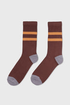 A pair of ribbed crew socks in a dark brown color, featuring two horizontal mustard yellow stripes near the cuff and contrasting light gray heels, toes, and soles, displayed against a light gray background.