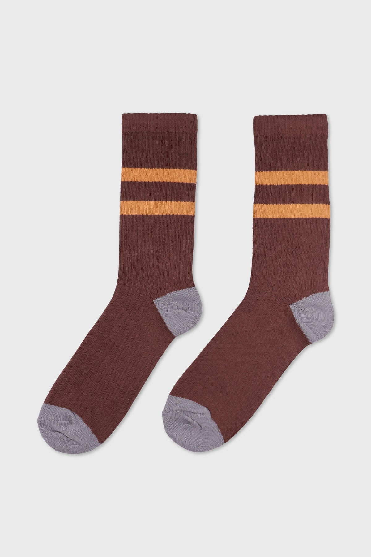 A pair of ribbed crew socks in a dark brown color, featuring two horizontal mustard yellow stripes near the cuff and contrasting light gray heels, toes, and soles, displayed against a light gray background.