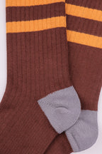 A close-up of a pair of ribbed knit socks in dark brown with two horizontal yellow stripes near the top and light gray heels and toes, shown on a plain white background.