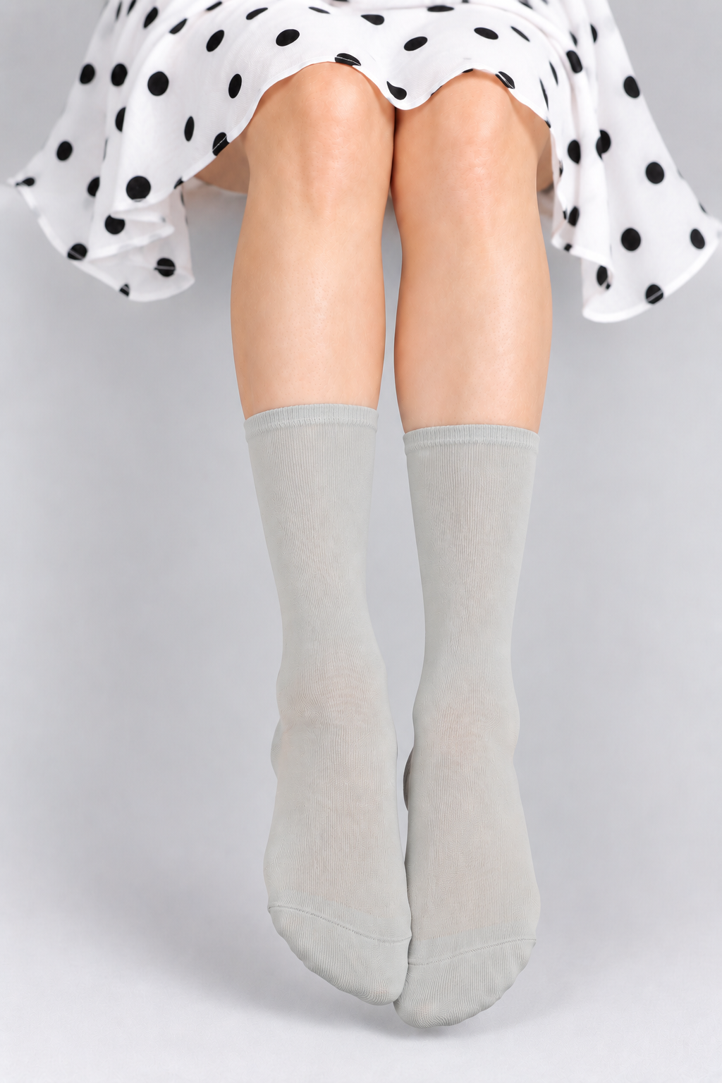 A person seated against a light gray studio background, wearing light gray knee-high socks and a white skirt with black polka dots. The product being shown is the socks.