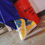 A person wearing bright blue corduroy pants and yellow low-top sneakers with colorful socks, sitting on a red metal barrel in an indoor setting.