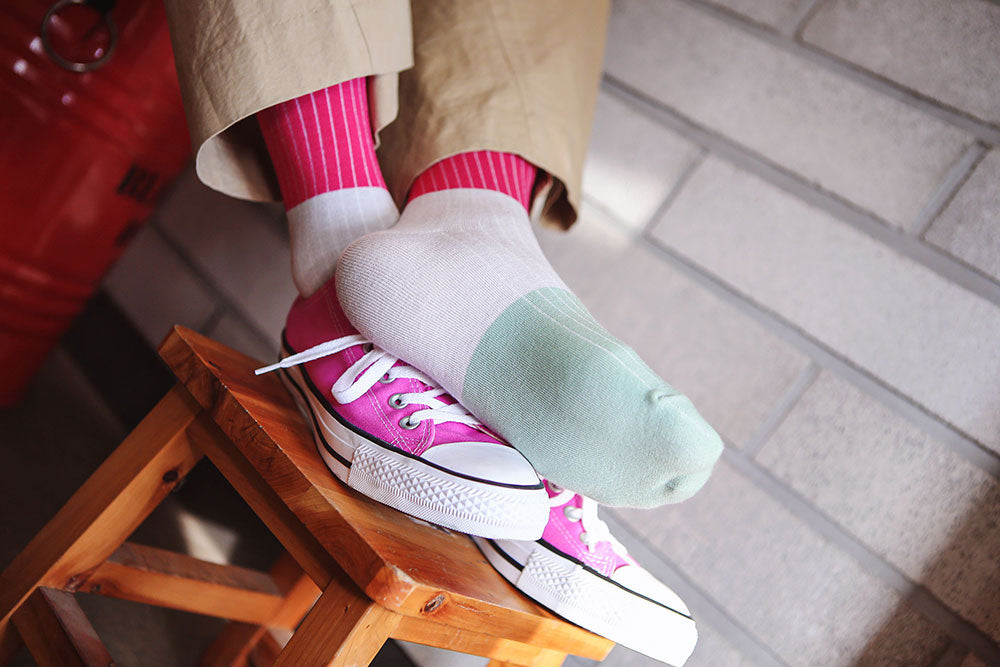 A close-up lifestyle shot of a person's feet resting on a wooden stool, showcasing a pair of multi-colored socks and pink sneakers. The socks feature a white ribbed top, a white midsection, and a mint green toe cap.