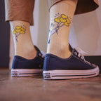 A person wearing navy blue Converse All Star sneakers and beige socks with a yellow flower and blue bird graphic, standing on a wooden floor.