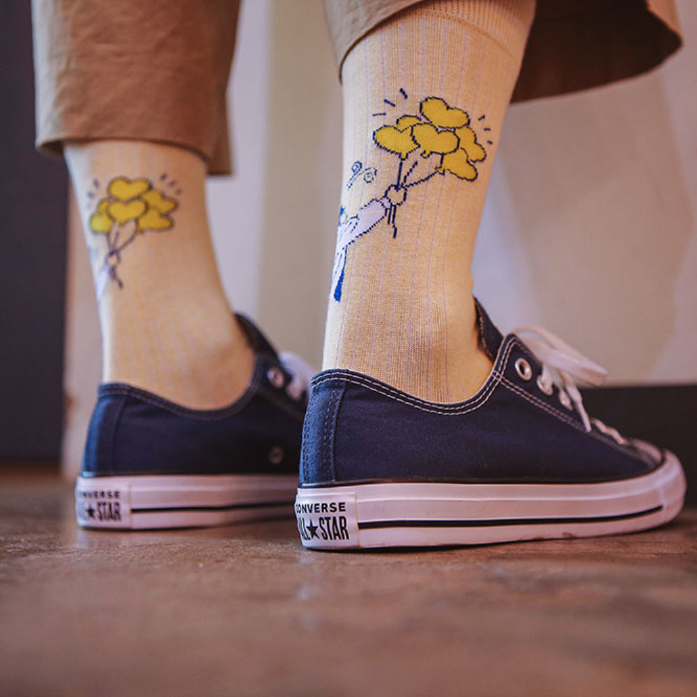 A person wearing navy blue Converse All Star sneakers and beige socks with a yellow flower and blue bird graphic, standing on a wooden floor.
