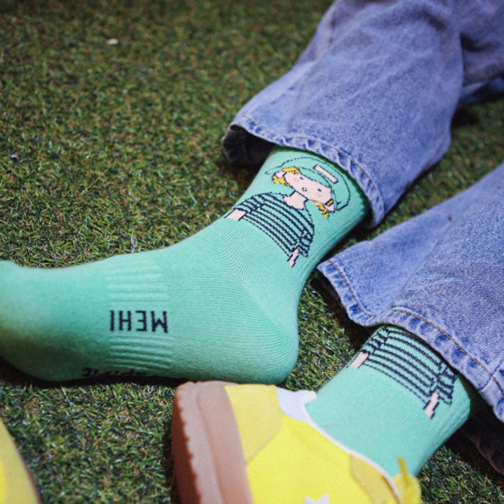A person wearing light green crew socks with cartoon graphics and black text, paired with blue jeans and yellow sneakers, lying on green artificial turf.