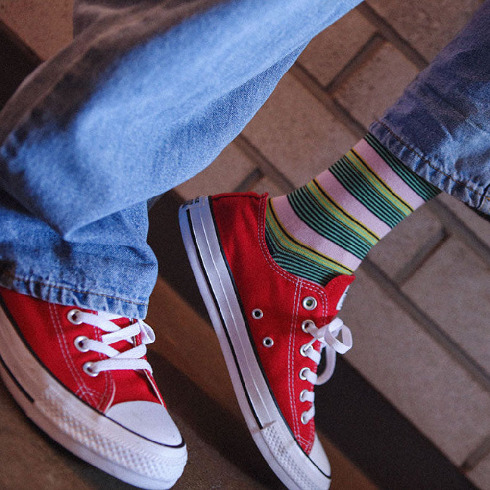 A close-up lifestyle shot of a person's legs crossed, wearing red canvas low-top sneakers with white laces, blue jeans, and a green, pink, and white striped sock, set against a brick floor background.