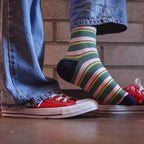A person wearing colorful striped crew socks with red low-top sneakers and blue jeans, standing on a wooden floor in front of a brick wall.