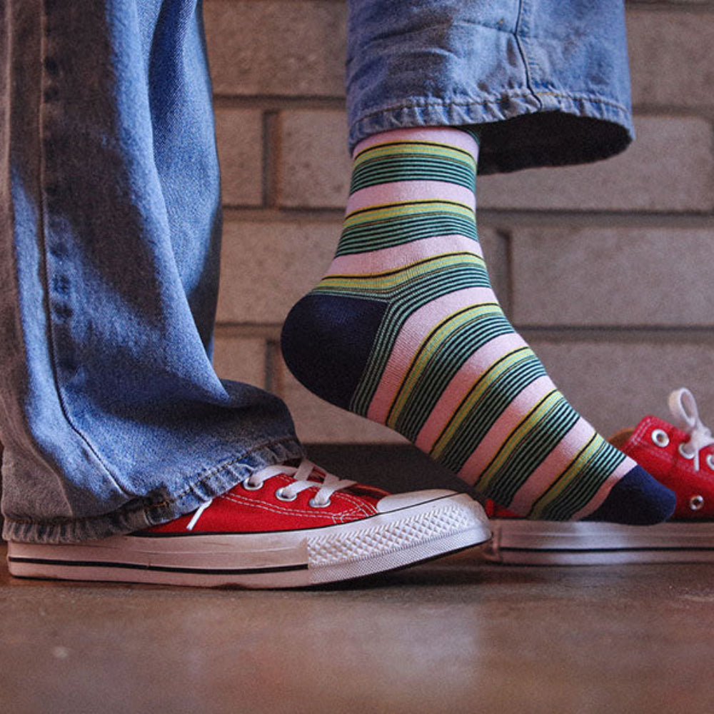 A person wearing colorful striped crew socks with red low-top sneakers and blue jeans, standing on a wooden floor in front of a brick wall.