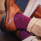 A close-up lifestyle shot of a person's legs crossed, wearing a brown leather brogue shoe, burgundy argyle socks, and beige trousers, resting on a dark leather chair.
