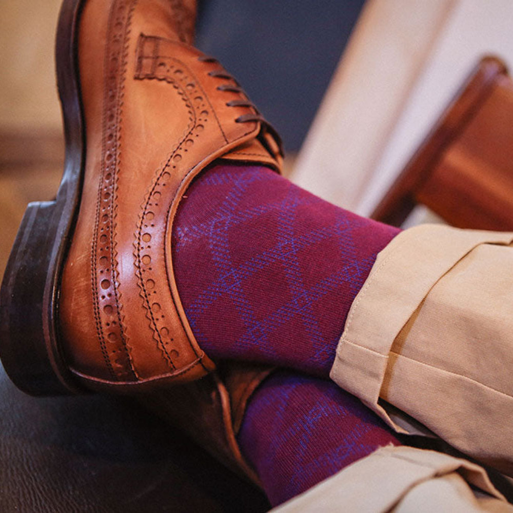 A close-up lifestyle shot of a person's legs crossed, wearing a brown leather brogue shoe, burgundy argyle socks, and beige trousers, resting on a dark leather chair.