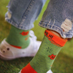Close-up of a person's legs wearing light blue ripped jeans, white sneakers, and green socks with orange cuffs and red cartoon cat graphics, standing on green grass.