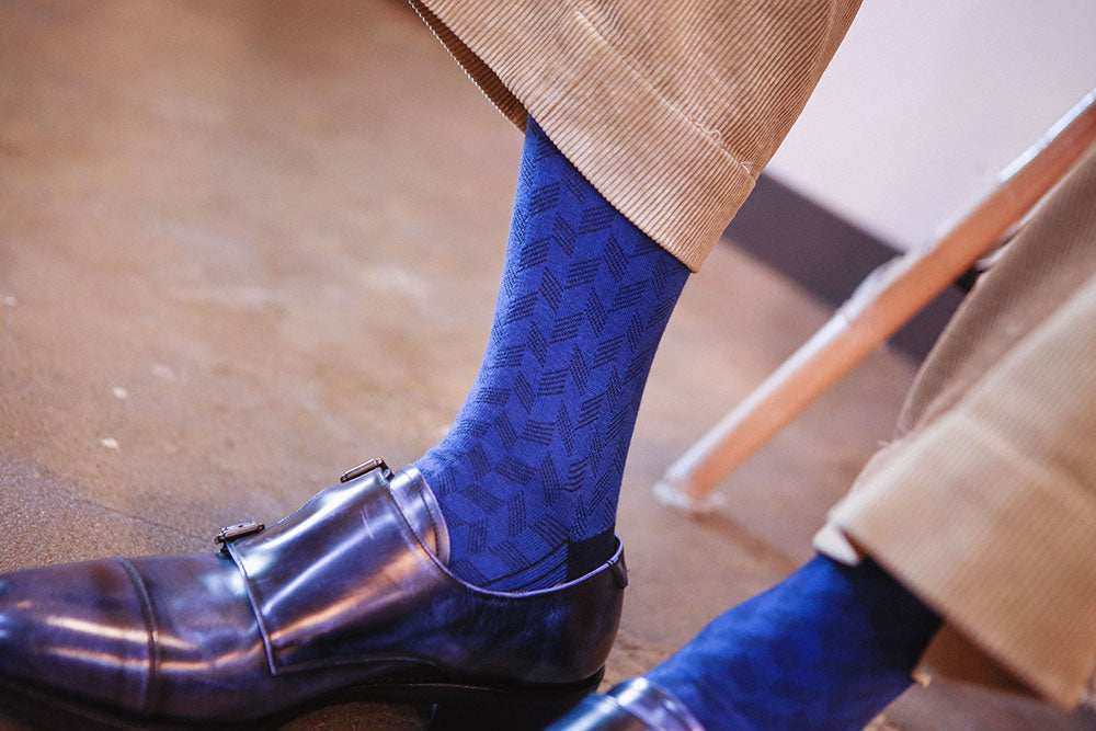 A close-up lifestyle shot of a person's legs wearing textured royal blue patterned socks with dark brown double-breasted loafers and beige trousers.