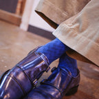 A close-up lifestyle shot of a person's feet wearing shiny dark blue patent leather double-breasted loafers, blue patterned socks, and beige corduroy trousers, standing on a polished brown floor.