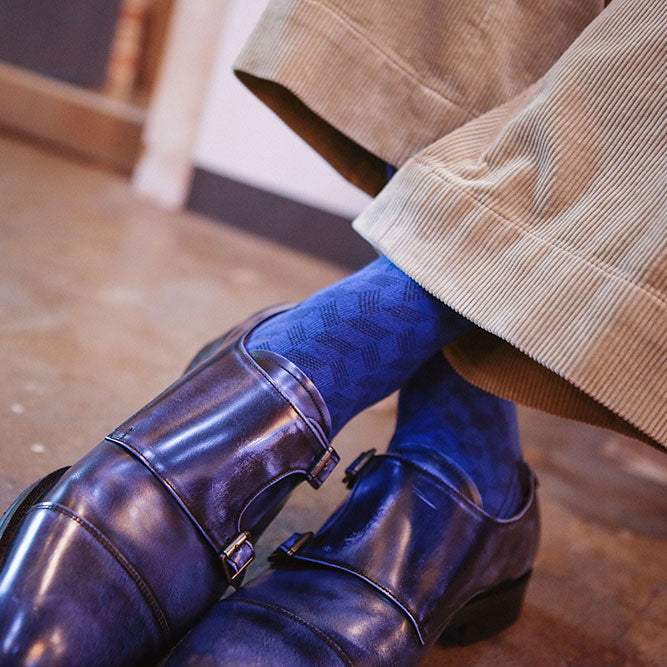 A close-up lifestyle shot of a person's feet wearing shiny dark blue patent leather double-breasted loafers, blue patterned socks, and beige corduroy trousers, standing on a polished brown floor.