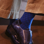 A close-up lifestyle shot of a person's legs and feet, featuring vibrant royal blue patterned socks paired with dark metallic purple leather shoes and brown corduroy trousers.