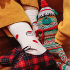 A close-up lifestyle shot of a person sitting on wooden steps, wearing white crew socks with red heart patterns and a Santa face, paired with a yellow sweater and red plaid shoes. Another leg wears a colorful knit holiday bootie sock with a green pine tree and red pom-pom.