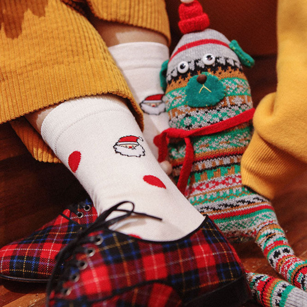 A close-up lifestyle shot of a person sitting on wooden steps, wearing white crew socks with red heart patterns and a Santa face, paired with a yellow sweater and red plaid shoes. Another leg wears a colorful knit holiday bootie sock with a green pine tree and red pom-pom.