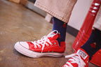Red low-top canvas sneakers with white laces and white rubber toe caps, worn with dark navy socks featuring a red Christmas tree pattern, shown on a person standing on a wooden floor.