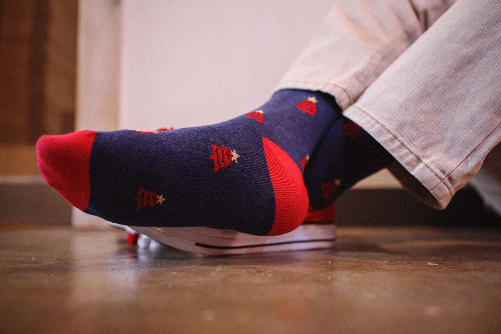 A close-up of a person's feet wearing navy blue socks with red toes, heels, and cuffs, decorated with small red Christmas trees and gold stars. The person is wearing light-colored jeans and standing on a wooden floor.