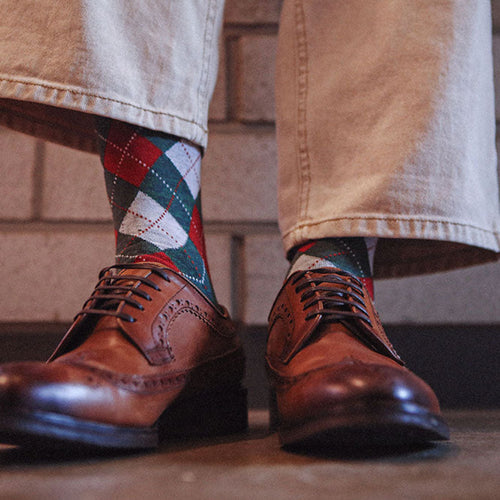 A close-up of a person's legs from the knees down, wearing beige jeans, colorful argyle socks, and brown leather wingtip oxford shoes against a brick wall background.