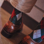 A close-up lifestyle shot of a person's legs and feet, showing brown leather oxford shoes paired with green, red, and white argyle socks and beige trousers, standing on a stone floor.