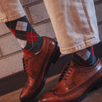 A close-up lifestyle shot of a person's legs wearing tan trousers, green and red argyle socks, and brown leather wingtip oxford shoes against a light brick wall background.