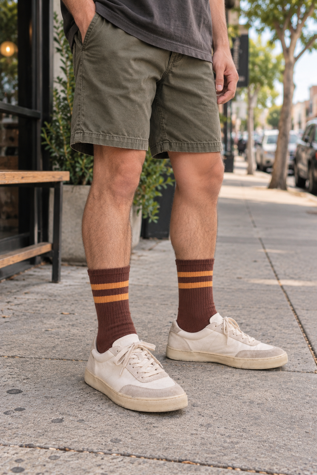 A person standing on a city sidewalk wearing olive green shorts, dark brown socks with thin orange stripes, cream sneakers, and a dark gray t-shirt. The background shows a blurred street scene with trees and parked cars.