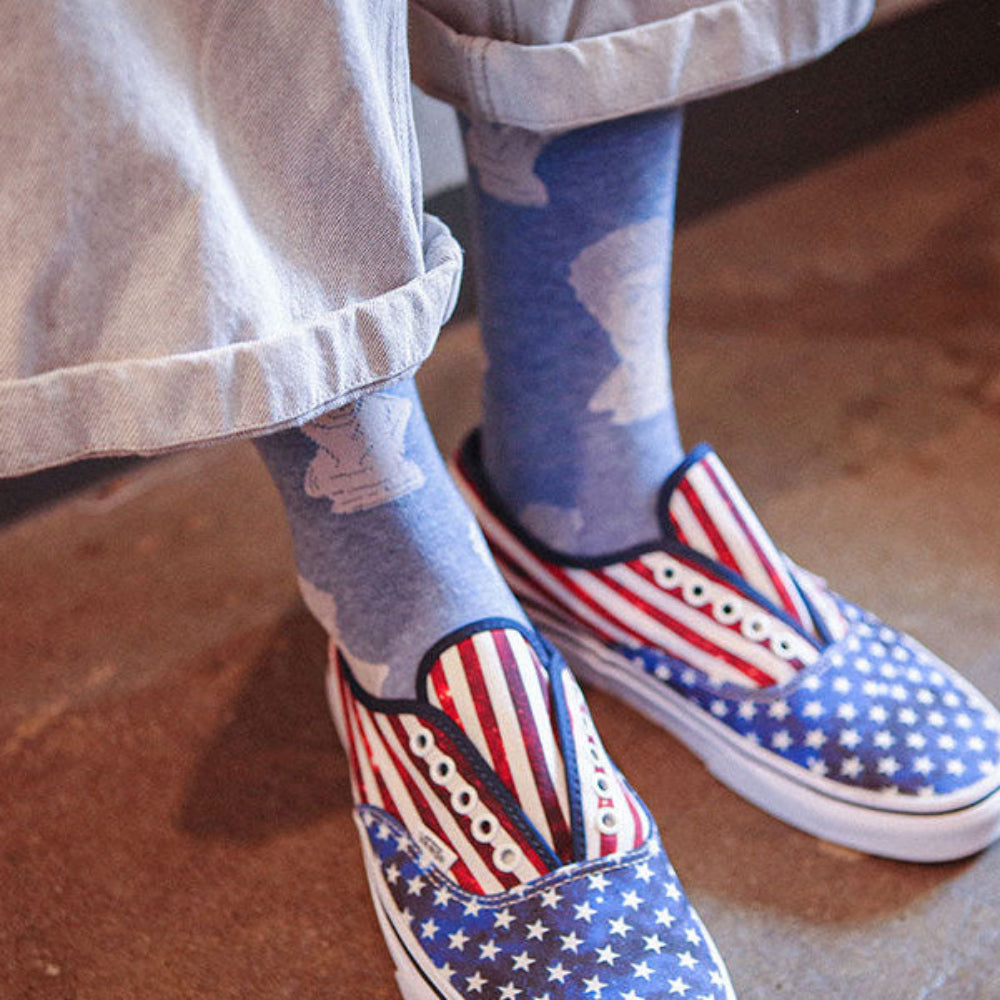A close-up of a person's feet wearing slip-on shoes with an American flag pattern, paired with blue socks and light gray cuffed pants, standing on a brown concrete floor.