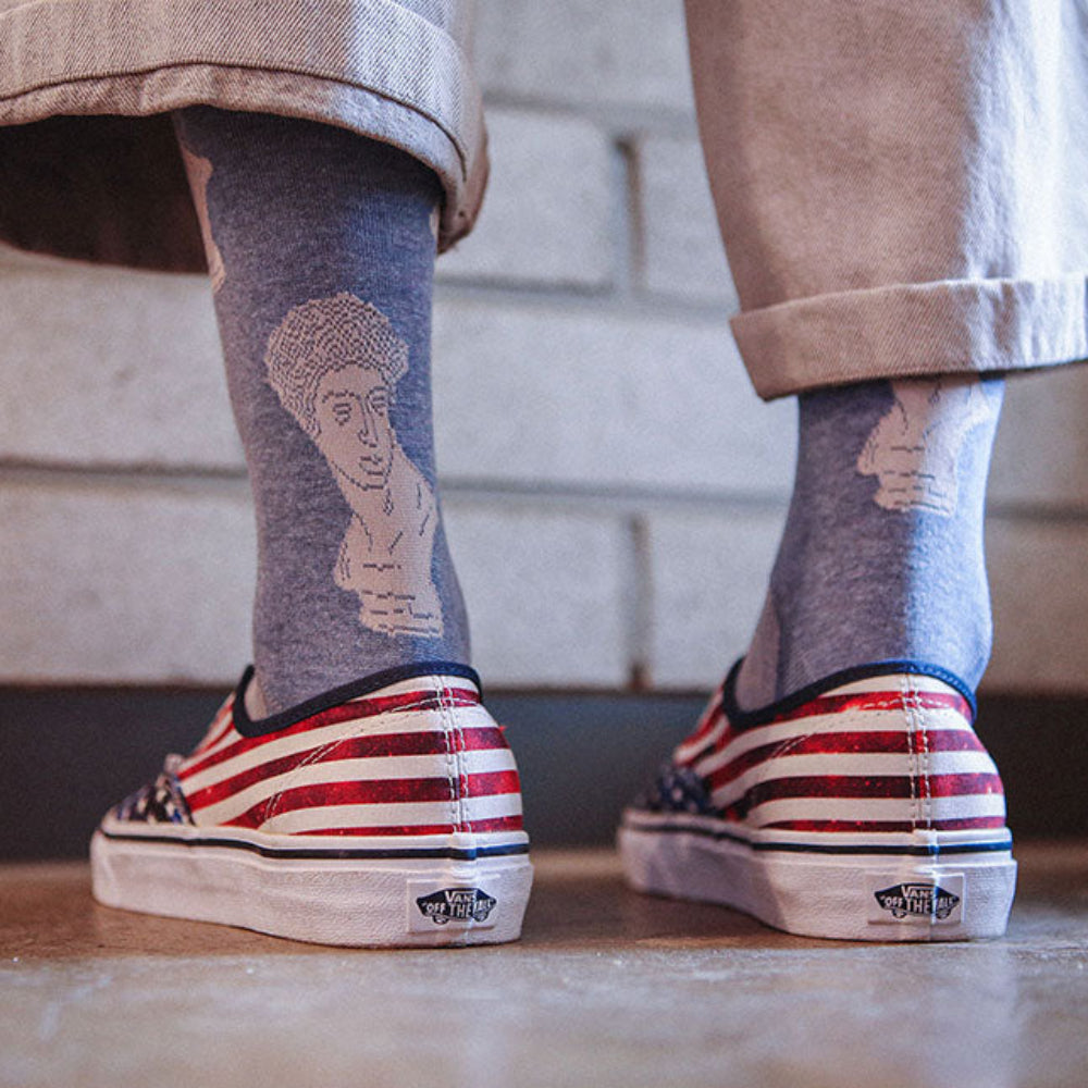 A person's legs wearing gray socks with white graphic prints and red, white, and blue striped Vans slip-on sneakers, standing on a wooden floor in front of a light brick wall.