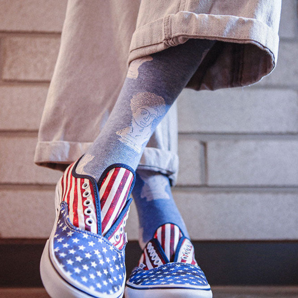 A person wearing slip-on slipper-style sneakers with an American flag pattern, paired with blue socks and light beige pants, standing against a light gray brick wall.
