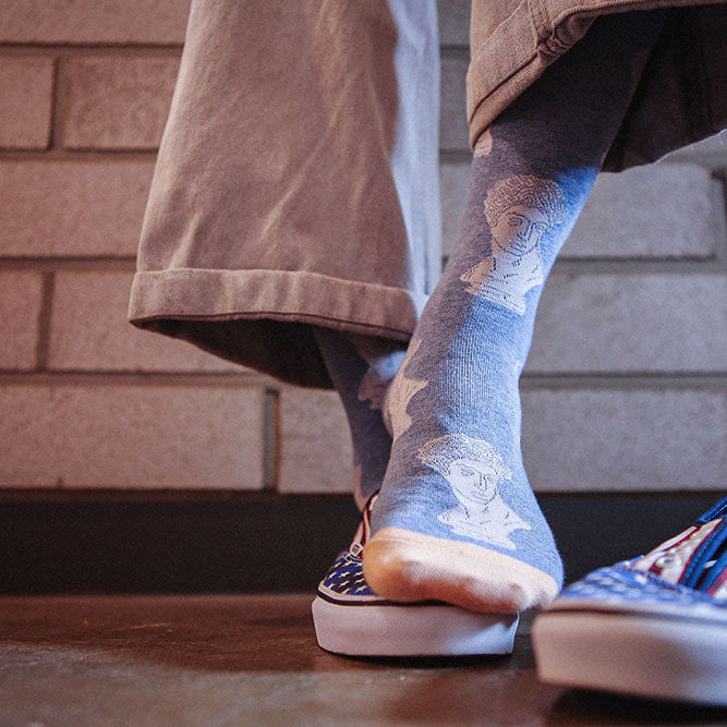 A person wearing light blue socks with a white cartoon bear design, paired with slip-on sneakers and cuffed khaki pants, standing in front of a brick wall.