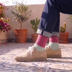 A person sitting on a tiled balcony wearing navy blue jeans with cuffed hems, pink and mint green striped socks, and beige suede sneakers. The background features potted plants and flowers under warm sunlight.