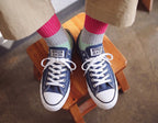 A person sitting on a wooden stool wearing navy blue Converse All Star low-top sneakers with white laces and toe caps, paired with pink and gray striped socks and khaki pants.