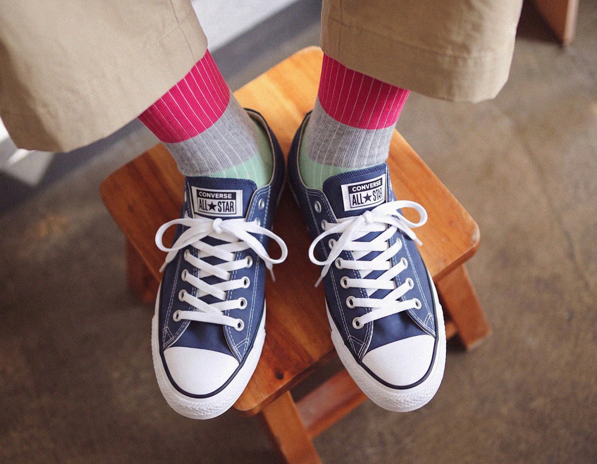 A person sitting on a wooden stool wearing navy blue Converse All Star low-top sneakers with white laces and toe caps, paired with pink and gray striped socks and khaki pants.