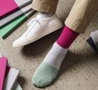 A person wearing beige pants and white sneakers, showing off a pair of color-block crew socks with pink and mint green sections and thin white pinstripes, standing on a speckled concrete floor with colorful notebooks nearby.